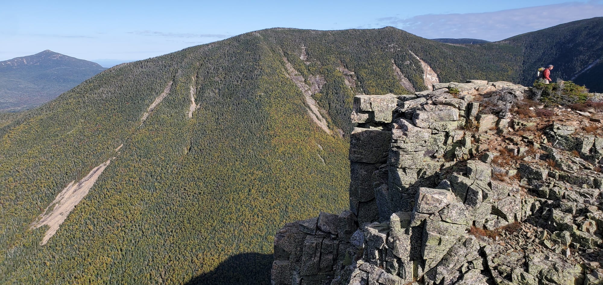 A hiker stands on the cliff edge of Bondcliff, with the slide-scarred flank of West Bond behind under a clear sky