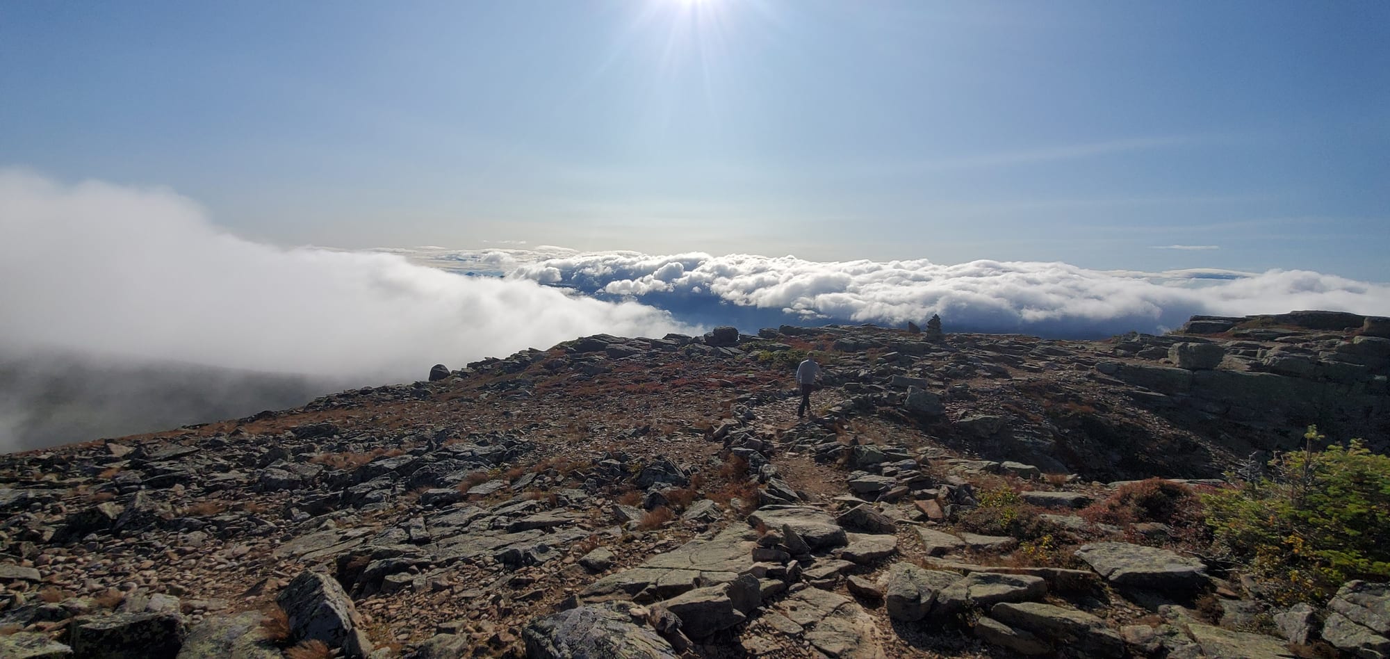 A hiker walking a rocky alpine ridge above a solid deck of cloud, with the sun low behind